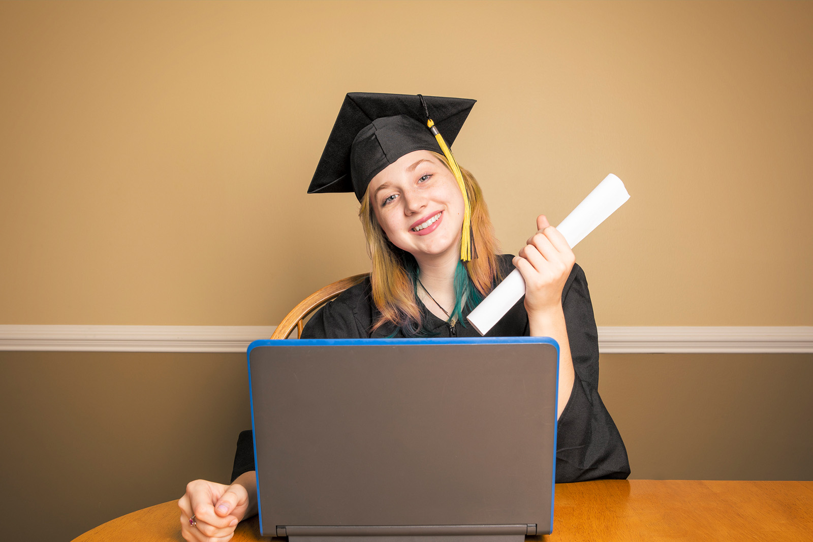 Young woman holding diploma using laptop, wearing graduation robes